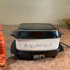 May include: A tall, slender, orange and black striped wooden cat figurine standing next to a rectangular white slow cooker with a black lid. The slow cooker features a blue floral vine pattern on the front and a black base with a control knob.