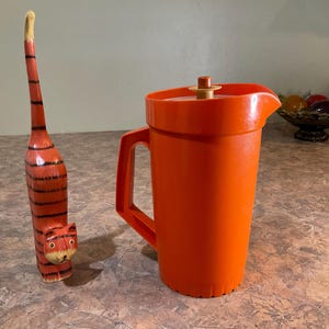 May include: A tall, slender, orange wooden cat figurine with black horizontal stripes and a long upright tail, positioned next to a bright orange plastic pitcher featuring a matching orange lid with a small central knob and a sturdy side handle.