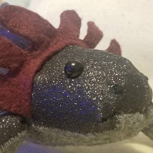 May include: A plush axolotl toy with a burgundy felt crown and a black button eye. The axolotl is made of a dark grey fabric with a glittery finish.