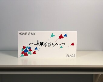 May include: White wooden sign with the text "Home is my happy place" with a black Mickey Mouse head in the middle of the word "happy". The sign is decorated with red, blue, and green Mickey Mouse head balloons.