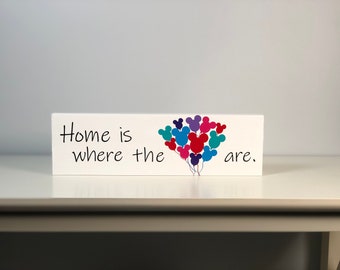 May include: White wooden sign with the text "Home is where the ___ are." with a cluster of colorful Mickey Mouse head balloons in the center.