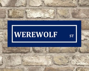 May include: A navy blue street sign with the text "WEREWOLF ST" in white lettering. The sign is framed in white and set against a brick wall background.