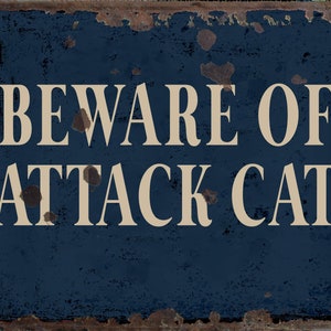 May include: A blue metal sign with white text that reads "Beware of Attack Cat". The sign has a distressed, rusty look.