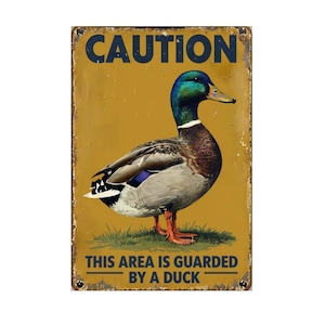 May include: A vintage-style metal sign with a yellow background and a detailed illustration of a mallard duck. The sign reads "CAUTION" at the top and "THIS AREA IS GUARDED BY A DUCK" at the bottom, with a distressed, aged appearance.