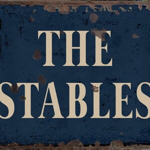 May include: A blue metal sign with rusty edges and white lettering that reads "THE STABLES".