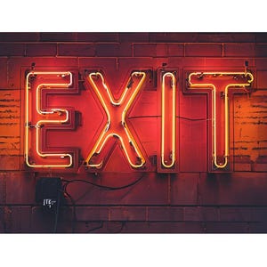 May include: Neon sign spelling out the word "EXIT" in bright orange against a dark red brick wall. The letters are illuminated with a warm glow, creating a striking contrast. The sign is mounted on a brick wall.