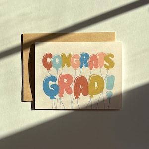 May include: A graduation greeting card with the words "Congrats Grad!" written in colorful balloon letters. The card is on a kraft paper background, with a matching envelope. The design is whimsical and celebratory.