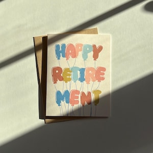May include: A greeting card with the words "HAPPY RETIREMENT" in colorful balloon letters. The card is on a beige background with a brown envelope. The image is lit by sunlight, creating shadows.
