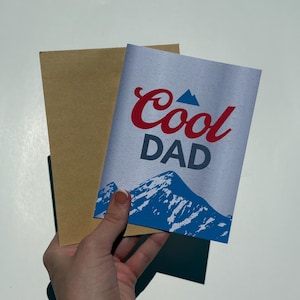 May include: A white greeting card featuring the red text Cool and the dark gray text DAD, accompanied by a blue mountain graphic at the bottom. The card is paired with a plain light brown envelope, presenting a simple and clean design aesthetic.