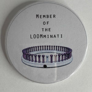 Könnte beinhalten: Ein weißer Knopf mit dem Text "MEMBER OF THE LOOMMINATI" und einer Grafik eines kreisförmigen Webstuhls mit lila und weißen Stiften.