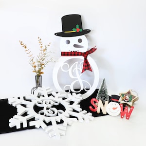 May include: White wooden snowman with a black hat and red and black plaid scarf. The snowman has a circle in the middle with a white monogram. There is a white snowflake with a monogram in the center, a small green pine tree, and a wooden sign that says "SNOW" with a snowman face.