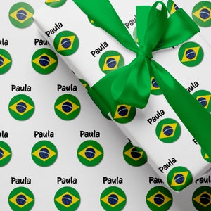 May include: White wrapping paper with a green ribbon and a repeating pattern of the Brazilian flag and the name "Paula".