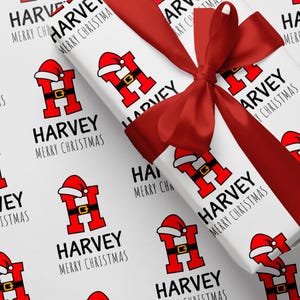 May include: White wrapping paper featuring a repeating pattern of a red letter "H" in a Santa suit, the name "HARVEY", and the words "MERRY CHRISTMAS" in black. A red satin ribbon is tied in a bow on top.