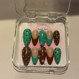 May include: A set of artificial nails in a clear plastic case. The nails feature various designs, including brown with green polka dots, solid tan, and green with tan polka dots. Some nails have pearl accents. The case reads "COLLECTION Nail Art".