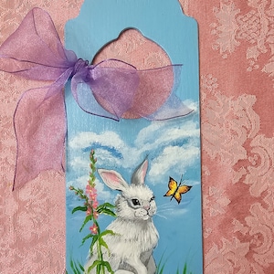 May include: A hand-painted wooden door hanger with a blue background, featuring a white rabbit sitting in a field of purple flowers and green grass. A yellow butterfly flies above the rabbit. The hanger has a hole at the top for hanging.