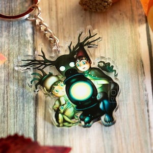 May include: A clear acrylic keychain featuring a cartoon image of two characters from the animated series, Over the Garden Wall, with a black lantern. The keychain has a silver chain and ring. The background includes fall foliage.
