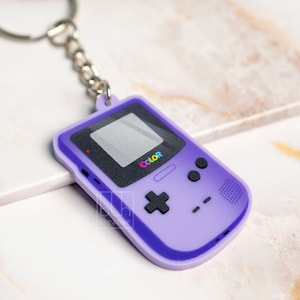Puede incluir: Llavero morado con forma de consola de juegos portátil retro con la palabra "COLOR" impresa en la pantalla.