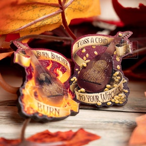 Op de afbeelding: Twee decoratieve, muntvormige houten ornamenten met een luitontwerp. De bovenste versiering is rood met gouden accenten en zegt "Toss a coin to your Witcher". De onderste versiering is rood met gouden accenten en zegt "Burn, Butcher, Burn".
