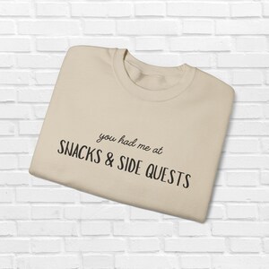 Może przedstawiać: Beżowa bluza z napisem "you had me at SNACKS & SIDE QUESTS" czarnym pismem. Bluza z okrągłym dekoltem jest złożona i leży na białej ceglanej ścianie.