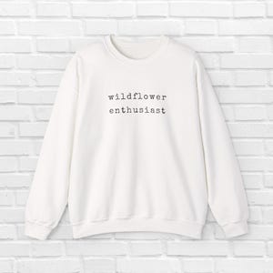 May include: White crew neck sweatshirt with the text "wildflower enthusiast" in black, centered on the front. The long-sleeved top is made of a soft material and is suitable for casual wear.