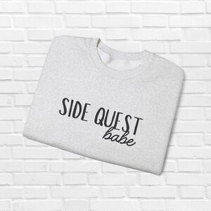 Może przedstawiać: Jasnoszara bluza z napisem "SIDE QUEST babe" w czarnym skrypcie. Złożona bluza znajduje się na tle białej ceglanej ściany. Bluza z okrągłym dekoltem wykonana jest z miękkiego materiału.