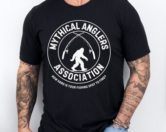 May include: Black t-shirt with a white circle graphic that says "Mythical Anglers Association" with a silhouette of Bigfoot holding fishing rods. The text below the circle says "How hard is your fishing spot to find?"