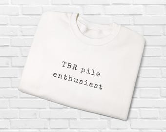 May include: A white crew neck sweatshirt with the text "TBR pile enthusiast" printed in black. The sweatshirt is folded and displayed against a white brick wall background. The text is in a simple, sans-serif font.