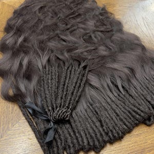 Peut inclure: Extensions de cheveux brun foncé, comprenant des mèches ondulées et des paquets de dreadlocks, disposées sur une surface en bois. Un ruban noir attache un paquet de dreadlocks. Les cheveux ont un aspect naturel et texturé.