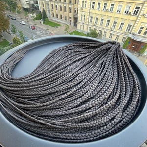 May include: Dark gray braided hair extensions, neatly arranged in a circular pattern. The braids are uniform in size and color, creating a sleek and stylish look. The hair is displayed on a dark gray surface.