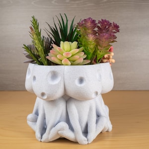 Puede incluir: Una maceta de cerámica blanca texturizada con forma de tres figuras, cada una con cabeza redonda y postura sentada. La maceta está llena de una variedad de suculentas artificiales en tonos verdes, rosas y morados.