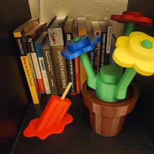 May include: A colorful flower arrangement with three flowers in a brown pot. The flowers are yellow, blue, and red. The pot is sitting on a shelf with books behind it. A red popsicle is melting on the shelf.