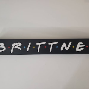以下が含まれることがあります： 黒い木製の看板に、白い文字で「Brittnee」という名前が書かれており、各文字の間にカラフルな点が付いています。