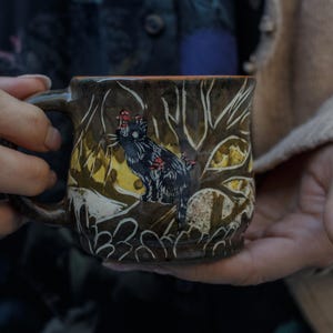 May include: A brown ceramic mug featuring a black cat and yellow mushrooms painted on it. The cat is perched on a tree branch against a brown and white background.