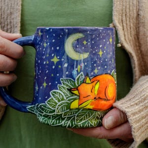 May include: A hand-painted ceramic mug. The mug has a dark blue night sky design with a crescent moon, stars, and a sleeping orange fox. The fox is nestled on green leaves, creating a charming, nature-inspired theme.