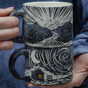 May include: A black and white ceramic mug with a hand-painted design of a mountain range and a crescent moon. The mug is stacked on top of another mug with a similar design.