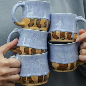 May include: A stack of four ceramic mugs with a light blue glaze. Each mug has a different design of brown and yellow socks on the bottom.