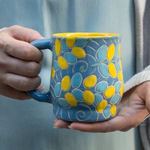Op de afbeelding: Een blauwe keramische mok met een geel en blauw bloemenmotief. De mok heeft een rond lichaam en een handvat.