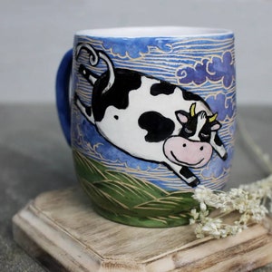 Könnte beinhalten: Eine Keramiktasse mit handbemaltem Design einer schwarz-weißen Kuh, die vor einem blauen und grünen Hintergrund durch die Luft fliegt.
