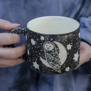 May include: A black ceramic mug with a white owl and crescent moon design. The mug is decorated with white stars and dots.