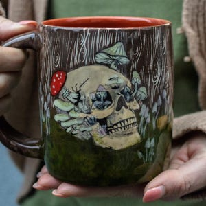 Könnte beinhalten: Keramikbecher mit dunkelbraunem Henkel und Rand, mit einem Totenkopf-Design mit Pilzen und einem rot gepunkteten Pilz. Der Becher hat einen Holzmaserung-Hintergrund und einen grünen Boden, was eine einzigartige und künstlerische Ästhetik schafft.