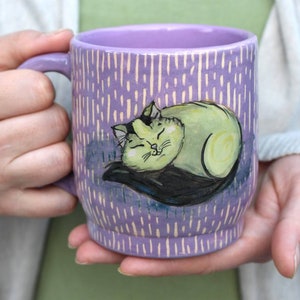 May include: A lavender ceramic mug with a sleeping cat illustration. The mug features a hand-painted design of a light green cat with black accents, resting on a dark blue pillow. The mug has a vertical striped pattern.
