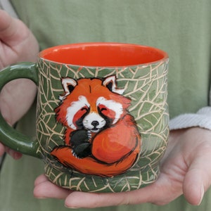 Peut inclure: Une tasse en céramique verte avec un bord orange présente un panda roux peint à la main enroulé sur un fond de feuilles vertes.
