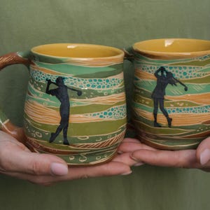 May include: Two ceramic mugs with a green, brown and yellow striped design. Each mug features a silhouette of a golfer in black.