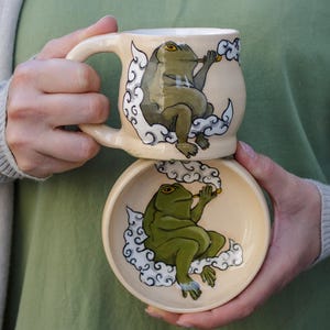 May include: Two ceramic mugs with frog illustrations. The top mug features a green frog with a pipe in its mouth, surrounded by clouds. The bottom mug features a similar green frog with a pipe in its mouth, surrounded by clouds.