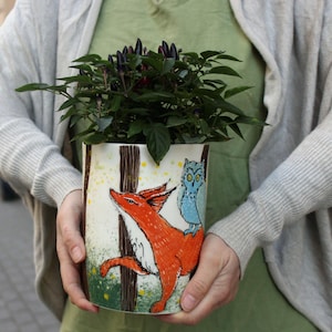 May include: A cylindrical white ceramic planter featuring a hand-painted illustration of a bright orange fox and a light blue owl perched on its back. The design includes brown tree trunks and green foliage. A small plant with dark purple peppers sits inside.