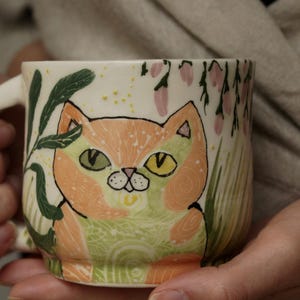 Puede incluir: Una taza de cerámica con un diseño de gato pintado a mano. El gato tiene pelaje naranja, ojos verdes y amarillos, y está rodeado de hojas verdes y flores rosas. La taza está sostenida por una persona.