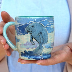 Peut inclure: Une tasse en céramique bleue et blanche avec une illustration peinte à la main d'un lamantin nageant dans les nuages. La tasse a un bord vert et une anse.