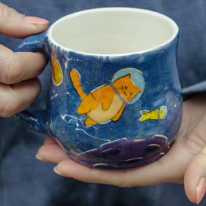 May include: A blue ceramic mug with a white interior and a space-themed design. The mug features a cartoon cat in a spacesuit floating in space with a yellow fish.