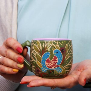 May include: A green ceramic mug with a pink interior and a painted design of two blue birds with red and yellow accents. The birds are surrounded by green leaves and flowers.