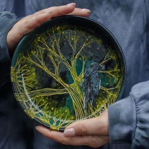 Könnte beinhalten: Eine schwarze Keramikschale mit handbemaltem Design eines Baumes mit gelben Ästen und einem schwarzen Raben. Die Schale hat einen dunkelgrünen Hintergrund.